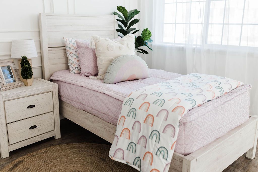 White twin bedframe with a textured euro pillow with tassels, pastel rainbow pillow, and a white blanket with ombre purple, orange and green rainbows