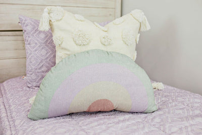 A close up view of a bed made with purple textured zipper bedding and cream euro pillow with tassels and a rainbow plush pillow.