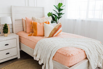 twin bed with Peach bedding with textured rectangle design with dark cream textured euro, orange, textured pillow with tassels, rainbow lumbar and knitted chenille throw with pom poms