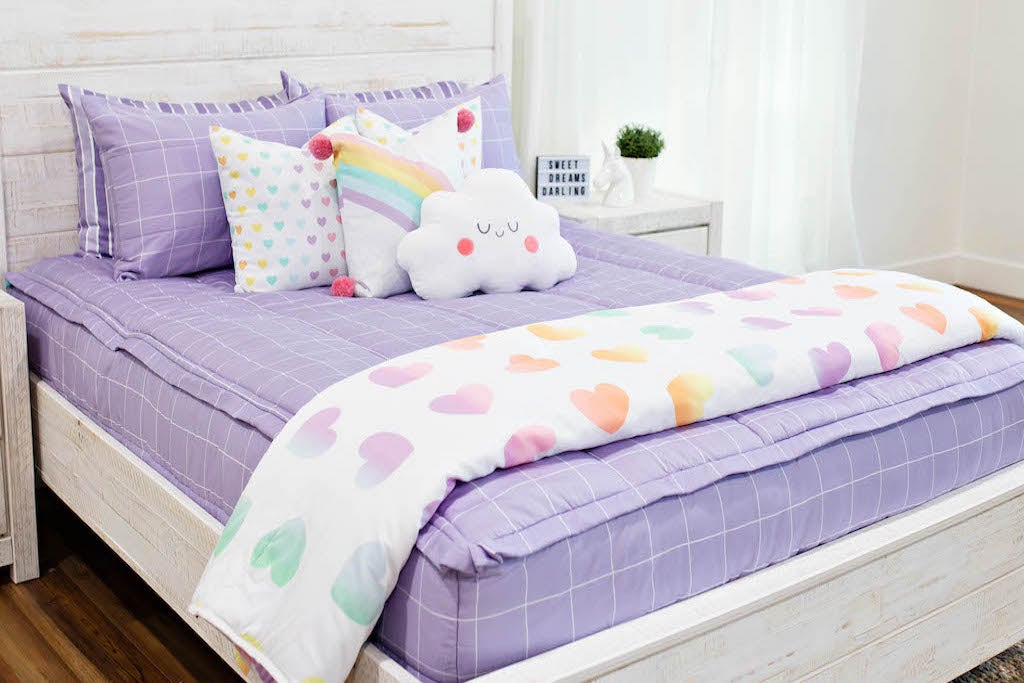queen bed with Purple bedding with white grid pattern, white pillow with rainbow ombre hearts, white pillow with rainbow and pink pom poms on the corner, plush marshmallow pillow, and white blanket with ombre hearts at the foot of the bed