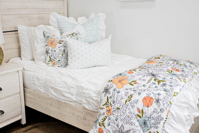 White twin bedframe with white textured bedding, white and blue striped euro with ruffle along the edge, floral printed pillow, white lumbar with small gray floral design and floral printed blanket with white ruffle along the edge