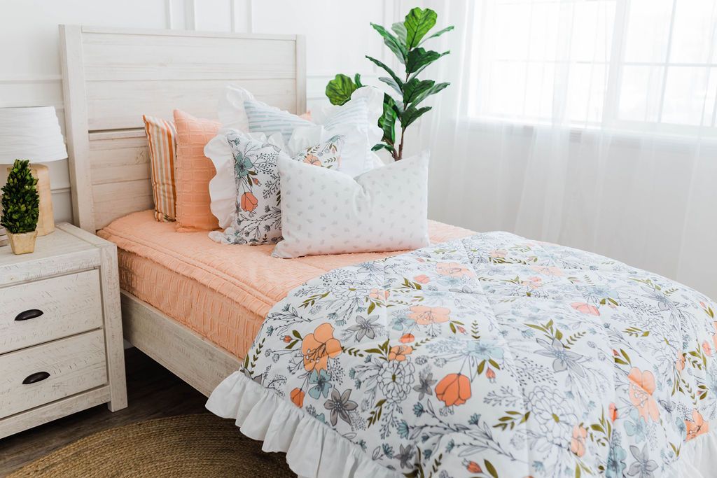 twin bed with Peach bedding with textured rectangle design with white and blue striped euro with ruffle along the edge, floral printed pillow, white lumbar with small gray floral design and floral printed blanket with white ruffle along the edge