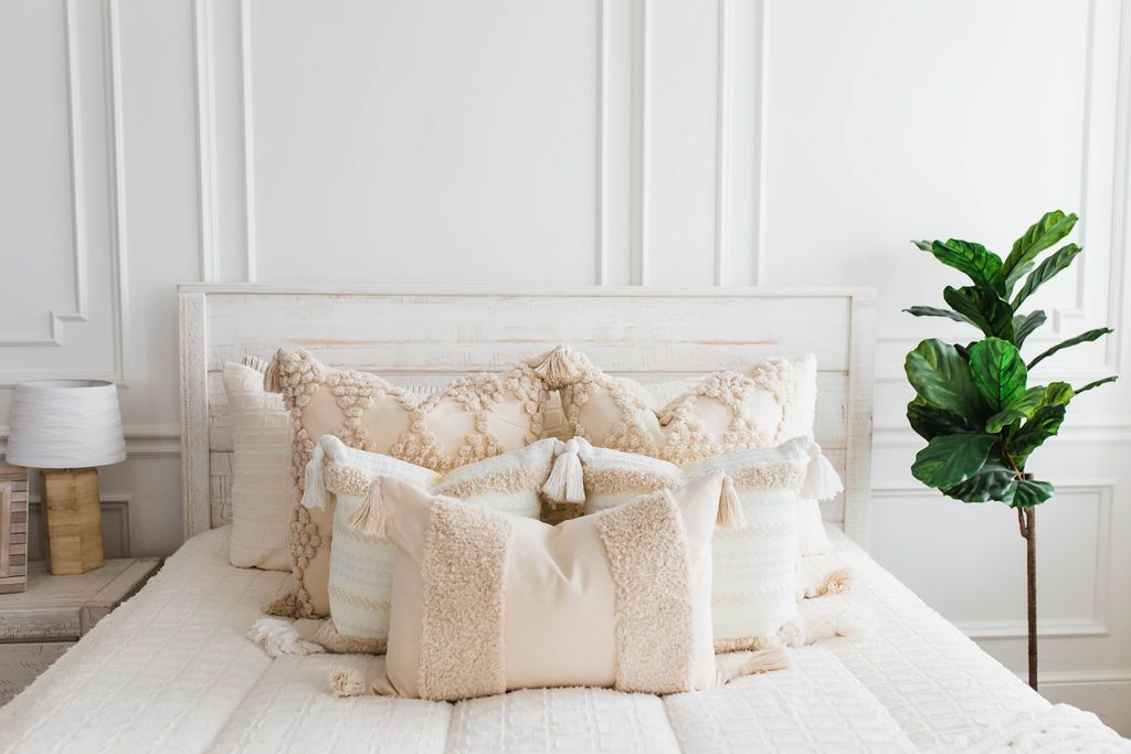 queen bed with Cream bedding with textured rectangle design and dark creamy textured euro, a cream and tan woven textured pillow and a textured dark creamy lumbar with tassels with an off white braided throw with pom poms along the edge