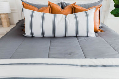 Gray zipper bedding with faux leather pillows, white XL lumbar with gray vertical stripes, and white blanket with horizontal gray stripes at the foot of the bed