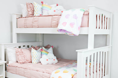 bunk bed with Blush pink bedding with cream textured euro pillows with tassels, one pastel rainbow pillow, and a white blanket with ombre purple, orange and green rainbows