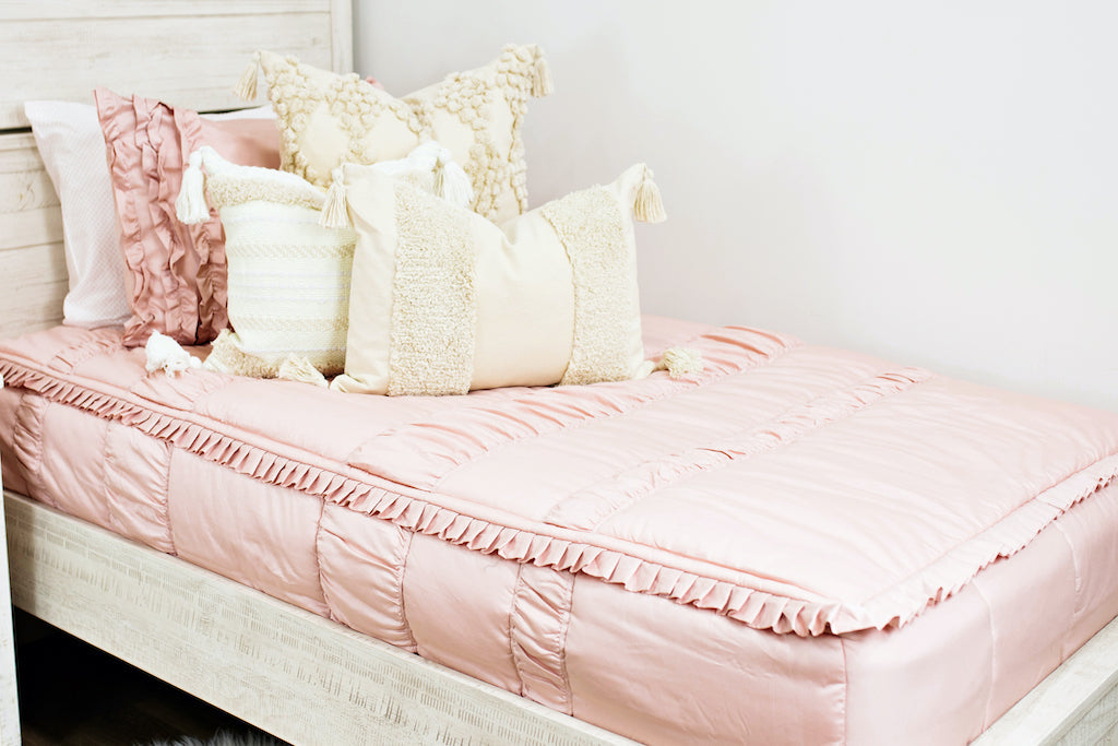 Twin bed with Blush pink bedding with dark creamy textured euro, a cream and tan woven textured pillow and a textured dark creamy lumbar with tassels 