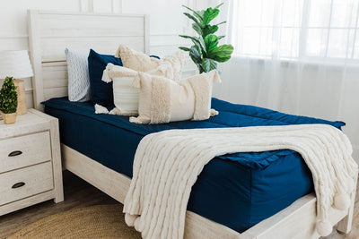 twin bed with Navy blue zipper bedding with dark creamy textured euros, cream and tan woven textured pillows and a textured dark creamy lumbar with tassels with an off white braided throw with pom poms along the edge