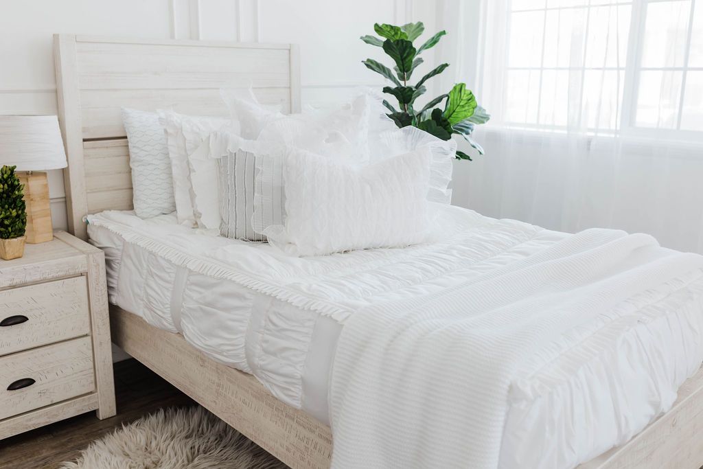 White twin bed frame with white textured bedding, one white ruffle polka dot euro, one ruffle gray/blue textured pillows, one white ruffle textured lumbar, and a textured white throw with braided tassels