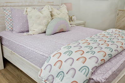 White queen bedframe purple textured bedding and textured euro pillow with tassels, pastel rainbow pillow, and a white blanket with ombre purple, orange and green rainbows