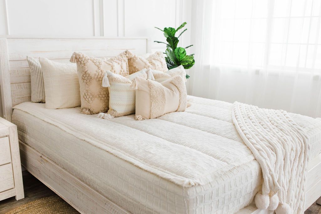 Queen bed with Cream bedding with textured rectangle design and dark creamy textured euro, a cream and tan woven textured pillow and a textured dark creamy lumbar with tassels with an off white braided throw with pom poms along the edge