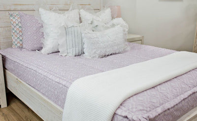 White queen bedframe with purple textured bedding, white ruffle polka dot euros, ruffle gray/blue textured pillows, white ruffle textured lumbar, and a textured white throw with braided tassels