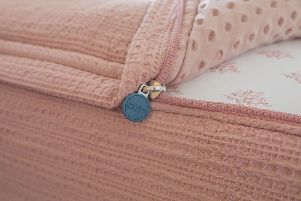 A close up view of a bed made with woven pink zipper bedding with the bed slightly unzipped to reveal the minky comforter and the floral pink and white pattern bottom cotton lining.