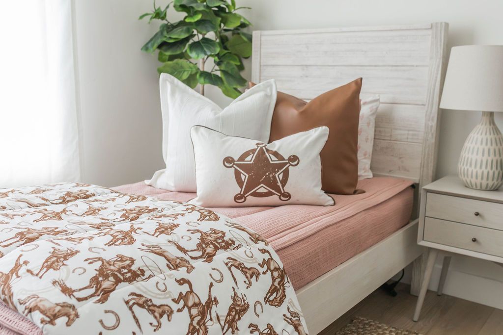A twin bed made with woven pink zipper bedding and an assortment of neutral and cowboy western print pillows and blanket.