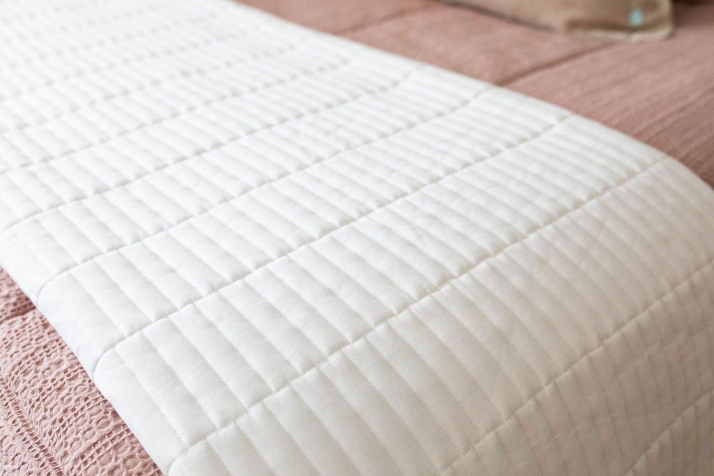 A close up of a bed made with woven pink zipper bedding and a white piano key quilted design blanket folded at the end of the bed.