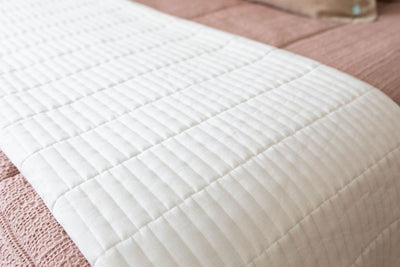 A close up of a bed made with woven pink zipper bedding and a white piano key quilted design blanket folded at the end of the bed.