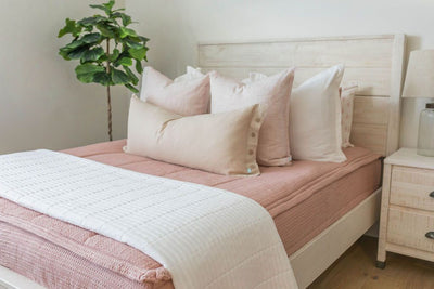 A queen bed made with pink woven textured zipper bedding and neutral and pink pillows and a white quilted blanket at the end of the bed.