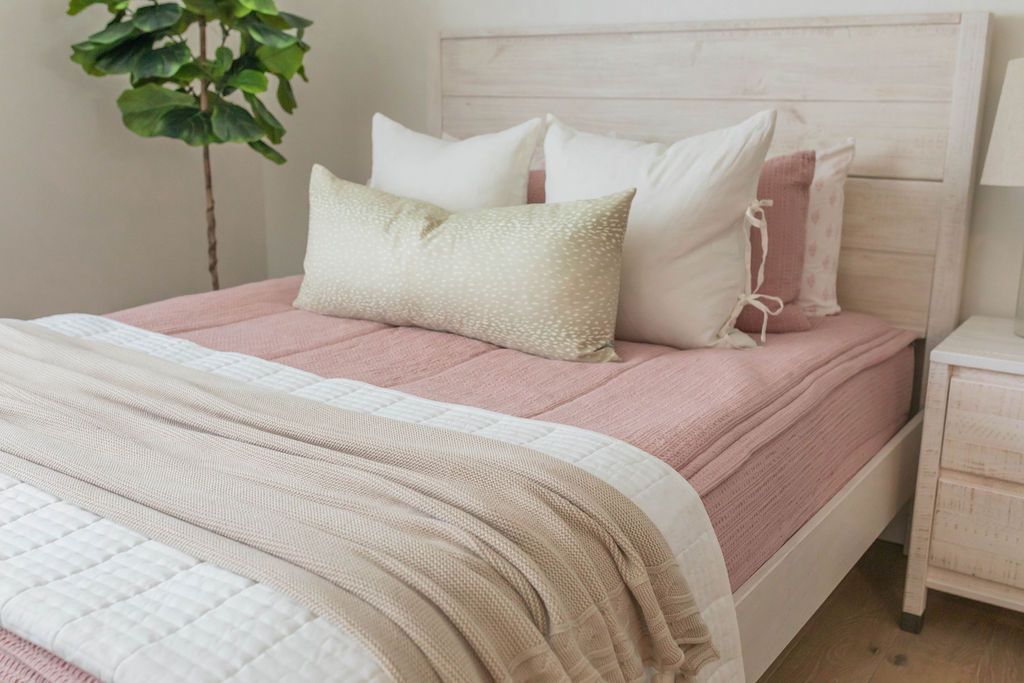 A queen bed made with pink woven textured zipper bedding and neutral cream pillows and matching blankets at the end of the bed.