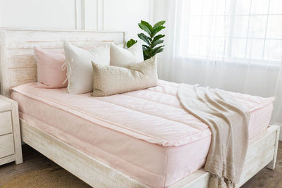 A brightly lit room with a bed made with a light pink zipper bedd and an assortment of neutral pillows and blanket.