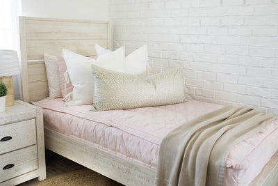 A twin bed made with light pink zipper bedding and cream and tan pillows and a woven blanket.