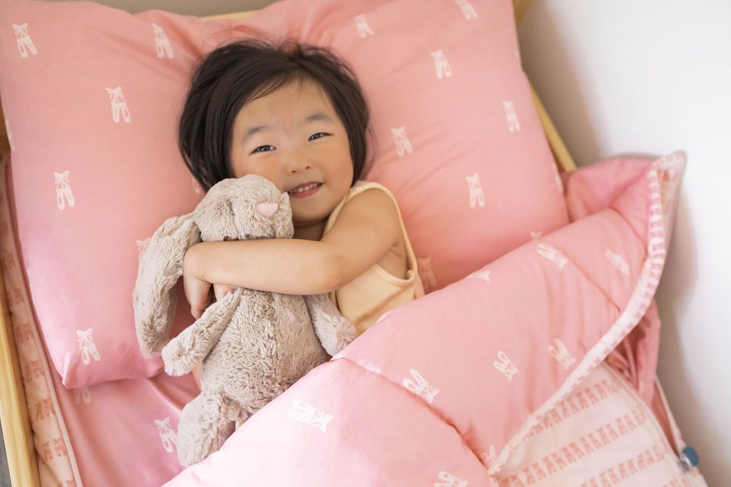 A young girl holding a stuffed animal bunny in a bed made with pink ballet shoes kids zipper bedding.