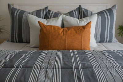 A close up of a bed made with light gray waffle textured zipper bedding and neutral and leather pillows and a matching striped blanket.