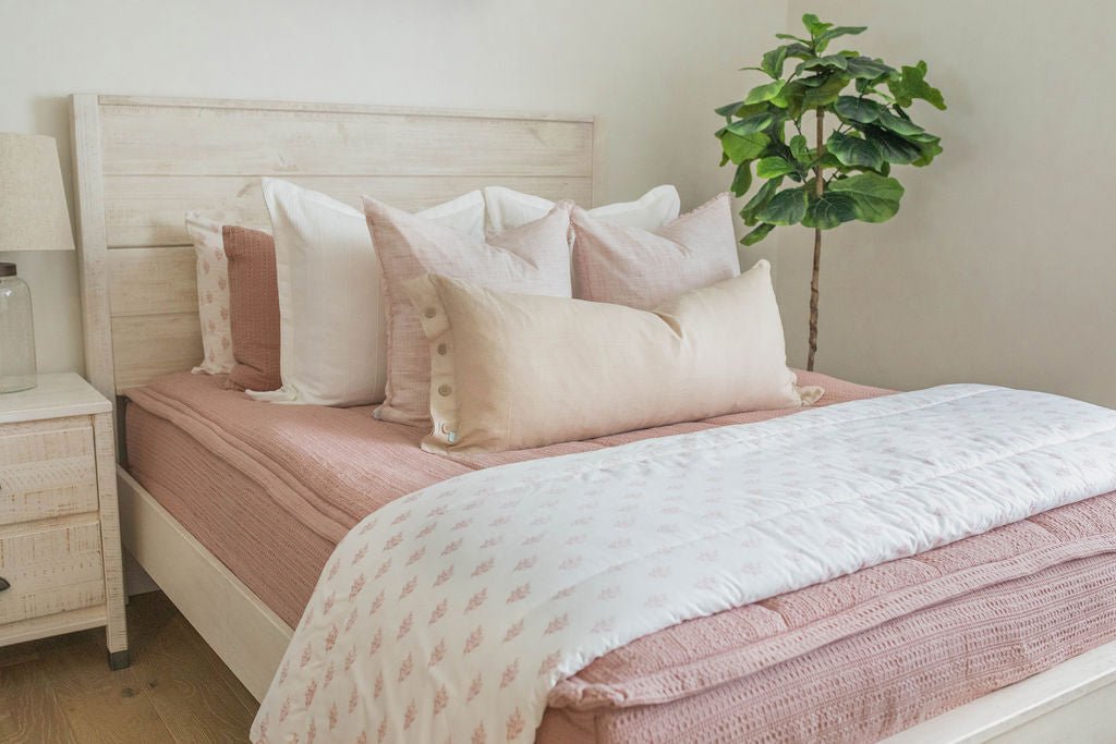 A queen bed made with woven pink zipper bedding and an assortment of neutral and light pink throw pillows and floral blanket.