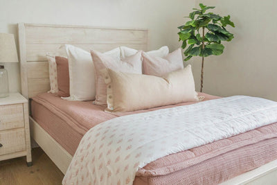 A queen bed made with woven pink zipper bedding and an assortment of neutral and light pink throw pillows and floral blanket.