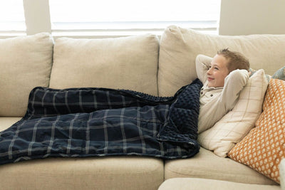 A boy smiling and laying on couch with a navy blue plaid children's blanket.