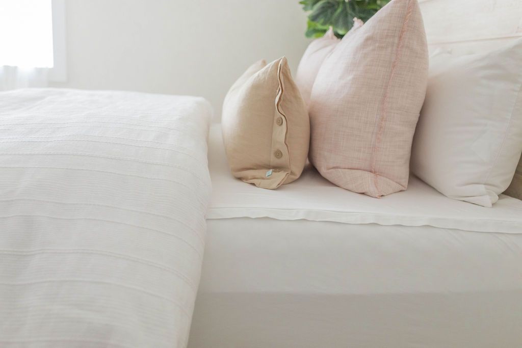 A close up sideview of a bed made with white zipper sheets and a textured white zipper duvet folded half way down the bed and adorned with neutral and light pink pillows.