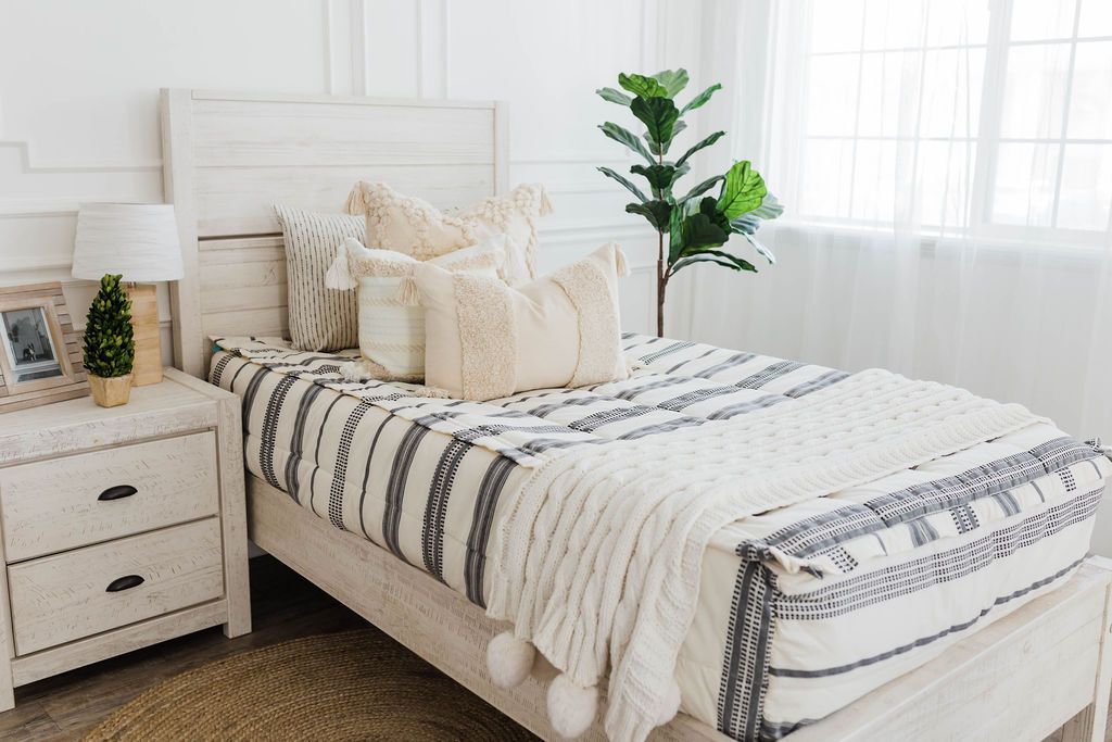 twin bed with cream and black woven striped bedding with dark creamy textured euro, a cream and tan woven textured pillow and a textured dark creamy lumbar with tassels with an off white braided throw with pom poms along the edge