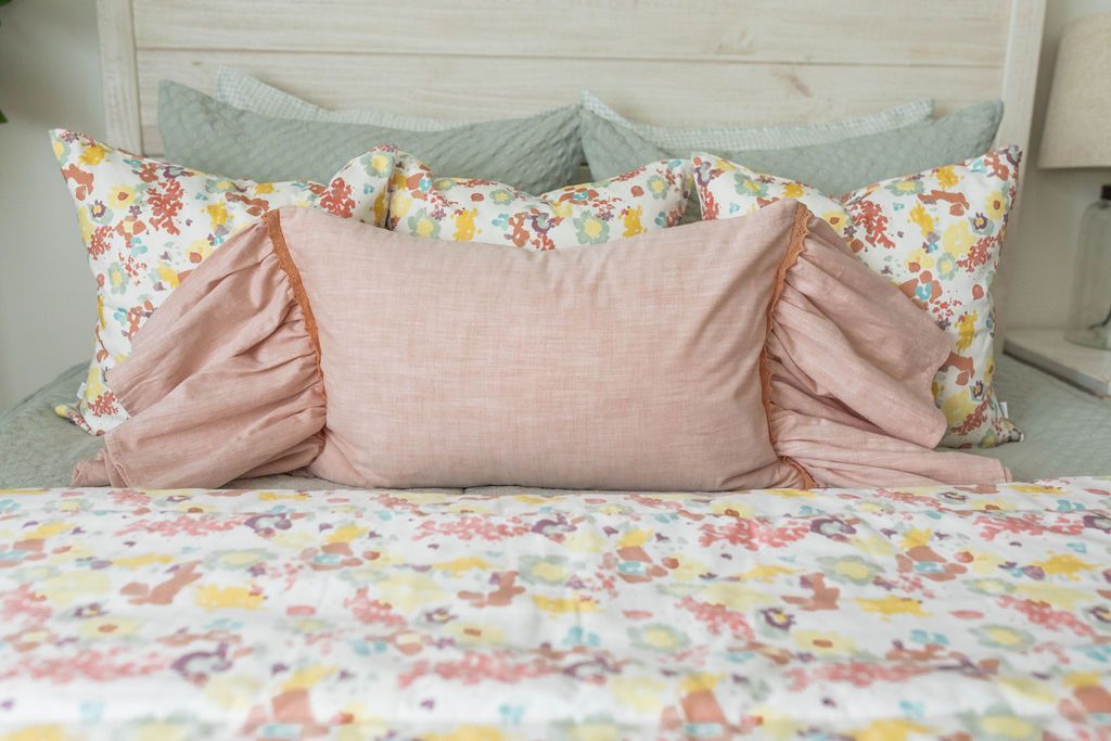 A close up of a bed made with sage green zipper bedding and colorful floral, pink, and green pillows and matching blanket.