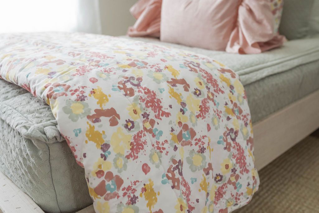 A close up side view of a bed made with sage green zipper bedding and a green, yellow, pink, and purple floral blanket folded at the end of the bed.