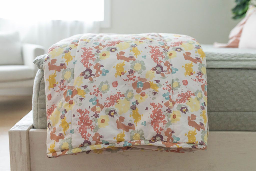 A close up side view of a bed made with sage green zipper bedding with a colorful floral blanket folded over the end of the bed.