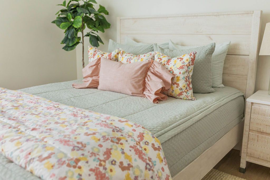 A queen bed made with sage green zipper bedding and floral pink and green pillows with a colorful matching blanket at the end of the bed.