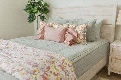 A queen bed made with sage green zipper bedding and floral pink and green pillows with a colorful matching blanket at the end of the bed.