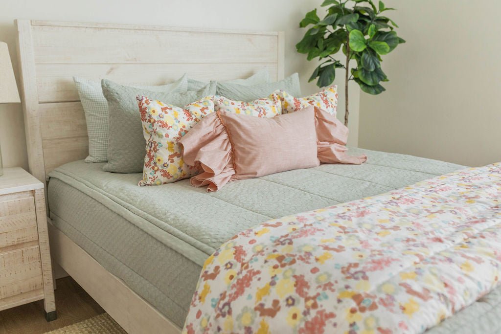 A bed made with soft green zipper bedding and an assortment of pink and multi-color floral pillows and blanket.