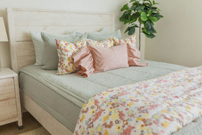 A bed made with soft green zipper bedding and an assortment of pink and multi-color floral pillows and blanket.