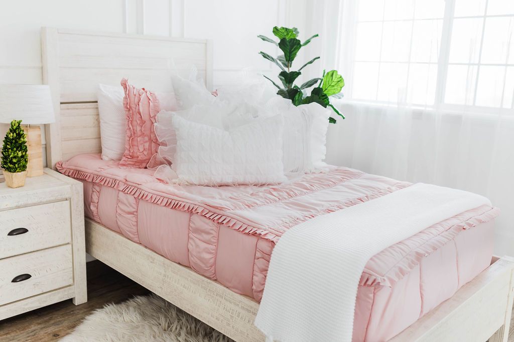 twin bed with Blush pink bedding with white ruffle polka dot euros,  ruffle gray/blue textured pillows, white ruffle textured lumbar, and a textured white throw with braided tassels
