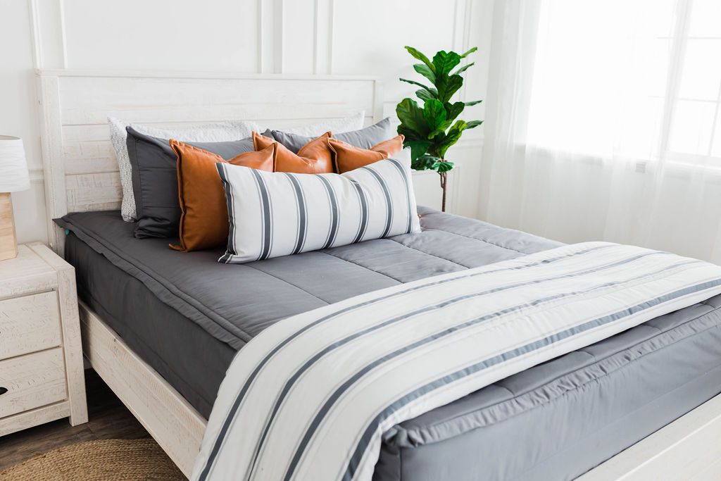 Queen bed with Gray zipper bedding with faux leather pillows, white XL lumbar with gray vertical stripes, and white blanket with horizontal gray stripes at the foot of the bed