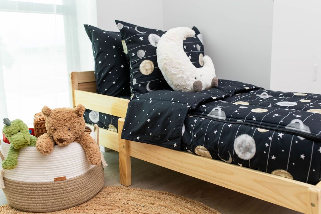 A toddler bed made with black planets and stars kids zipper bedding and a basket of stuffed animals beside it.