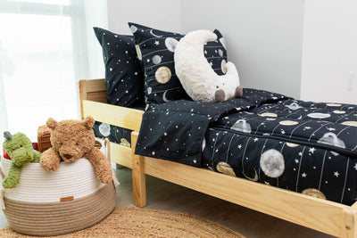A toddler bed made with black planets and stars kids zipper bedding and a basket of stuffed animals beside it.