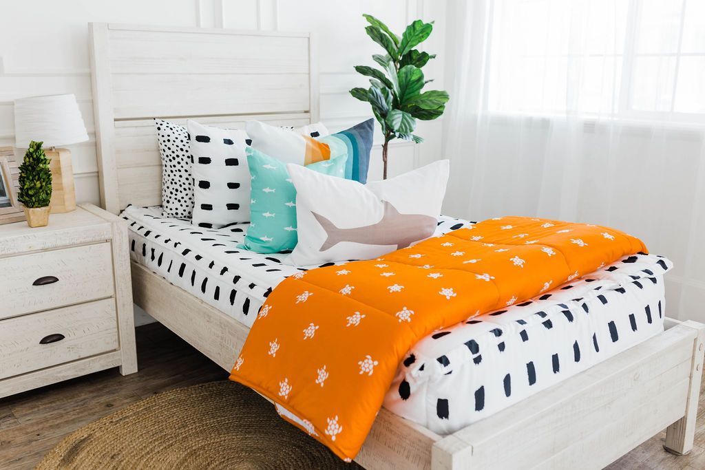 twin bed with White bedding with black dashed lines and white, teal, orange, white striped euro, teal pillow with white shark print, white lumbar with gray shark print, and orange blanket with white turtle print at the foot of the bed