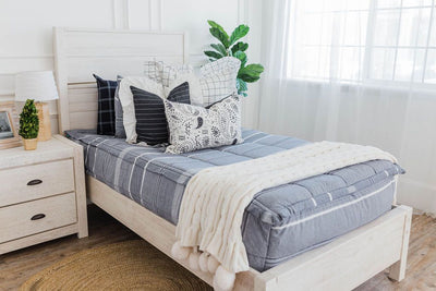 twin bed with Deep navy woven stripe bedding cream and black grid euros with ruffle along the edge, charcoal striped pillow with white ruffle along the edge, cream lumbar with charcoal paisley print, and cream textured blanket with pom poms