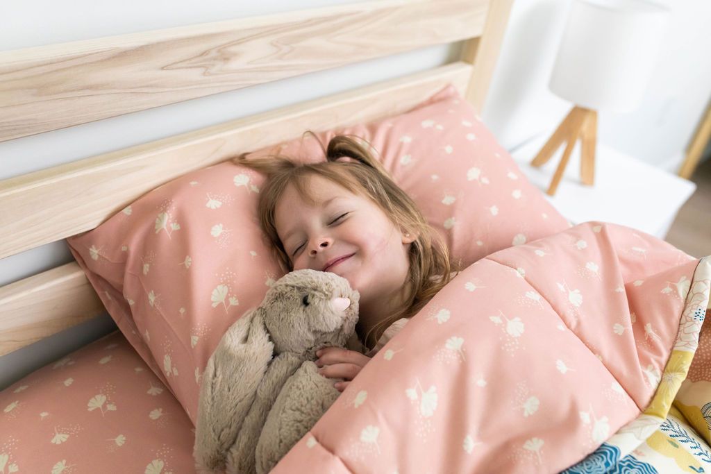 A young girl holding a stuffed animal sleeping in a bed made with yellow, pink, and blue floral kids zipper bedding.