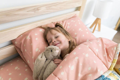 A young girl holding a stuffed animal sleeping in a bed made with yellow, pink, and blue floral kids zipper bedding.