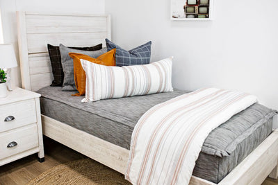 A twin bed made with a deep brown gray zipper bedding and striped neutral pillows and a coordinating blanket.