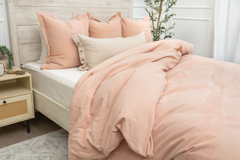 A brightly lit bedroom with a bed made with white zipper sheets and a peach zipper duvet folded partially half way down the bed and adorned with matching peach and neutral pillows.