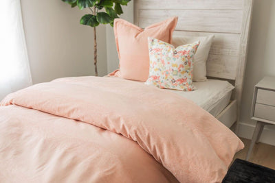 A twin bed made with white zipper sheets and a peach zipper duvet folded half way down the bed and adorned with a peach and multi-color floral pillows.