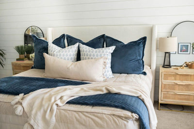 A bed made with tan faded stripes zipper bedding and an assortment of neutral and blue velvet pattern throw pillows and matching blankets.