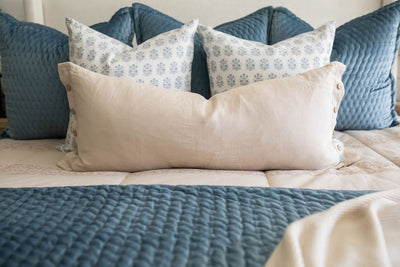 A close up view of a bed made with tan zipper bedding and an assortment of neutral and blue velvet throw pillows and blankets.